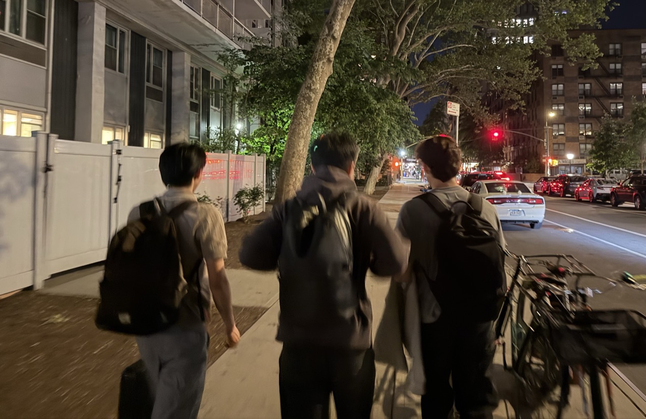 Three friends walking down a New York City street at night, seen from behind, wearing backpacks.